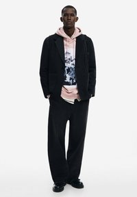 Blazer negro con costuras visibles, superpuesto a una sudadera rosa con un gráfico. Combinado con pantalones negros holgados y zapatos negros gruesos.