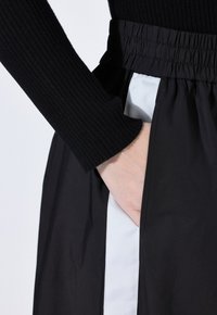 Main dans la poche latérale d'un pantalon noir avec une rayure verticale blanche et une taille élastique, portant un haut noir à manches longues en côtes.