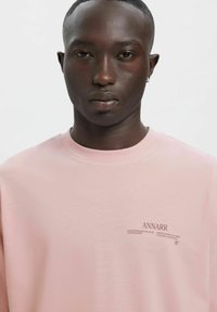 Różowy bawełniany T-shirt z okrągłym dekoltem i krótkimi rękawami. Zawiera nadrukowany tekst po lewej stronie, "ANNARR" oraz inne detale w ciemnej czcionce.