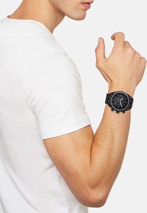 Sort chronograf ur med en tekstureret skive og tre underdials, forsynet med et armbånd i rustfrit stål, båret på et håndled med lys hudtone.