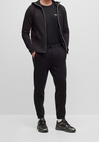 Man i svart zip-up hoodie, svart t-shirt, svarta joggers, vita strumpor och svarta träningsskor står med händerna i fickorna.