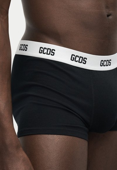 Κοντινό πλάνο ενός ατόμου που φοράει μαύρα μποξεράκια με λευκή μέση που φέρει επαναλαμβανόμενο λογότυπο "GCDS" με μαύρα γράμματα.