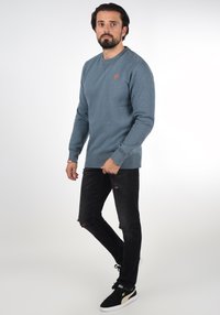 Blå sweatshirt med ribbade muddar och halsringning, med en liten orange logotyp. Bärs med svarta trasiga jeans och svarta sneakers med gulddetaljer.