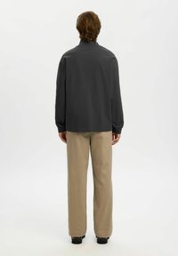 Donkergrijze lange mouw shirt met hoge kraag, gecombineerd met lichtbeige wijde broeken en zwarte schoenen, gezien van achteren.