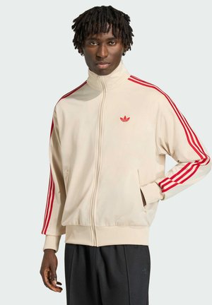 Mann trägt cremeweiße Adidas-Trainingsjacke mit roten Streifen und schwarze Hose, steht mit linker Hand in der Tasche vor einfachem Hintergrund.