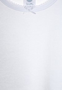 White cotton shirt with a ribbed texture, featuring a scalloped neckline and a small silver ribbon bow at the center. Label visible at the top.