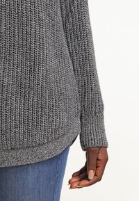 Grijze gebreide trui met een gestructureerd ribpatroon, voorzien van lange mouwen en een ronde zoom. Gemaakt in combinatie met blauwe denim jeans.