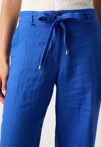 Blue linen pants with a drawstring waistband and metal toggles. Texture is smooth and slightly wrinkled, featuring side pockets.