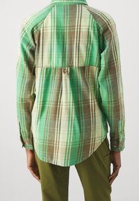 Zelená a hnědá kostkovaná košile s knoflíkovým zapínáním vzadu, dlouhými rukávy a zaobleným lemem. Materiál působí měkce s mírně kartáčovanou texturou.