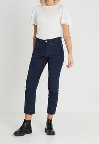 Pantalon en velours côtelé bleu marine, coupe slim et longueur cheville. Associé à un t-shirt à manches courtes blanc cassé et des bottes noires à lacets.