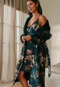 Woman standing in a floral nightdress and matching robe, in a softly lit bedroom with sheer curtains and neutral decor.