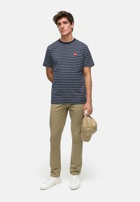 Marine stripete T-skjorte med en liten rød logo, khaki bukser, hvite joggesko, og en beige caps holdt i den ene hånden, stående mot en ensfarget bakgrunn.