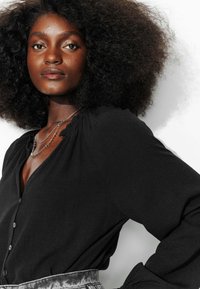 Blouse noire à manches longues avec un col en V et fermeture à boutons, présentant des épaules froncées et un tissu texturé. Des colliers en argent superposés sont portés.