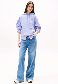 Lila oversized hoodie met voorzak, gedragen over een lichtpaarse geruite blouse. Wijde blauwe jeans en witte sneakers maken de outfit compleet.