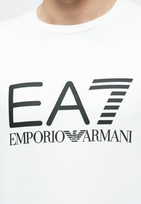 Fehér póló fekete EA7 Emporio Armani logóval és stilizált sas emblémával, középen a mellkason.