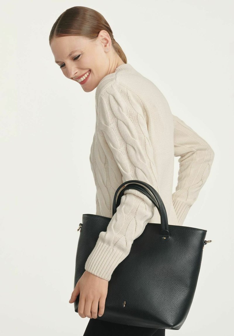 Czarna skórzana torebka o teksturowanym wykończeniu, z dwoma okrągłymi uchwytami i złotymi akcentami. Modelka nosi kremowy sweter w sploty, pozując z boku.