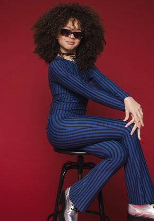 Blauwe en zwarte gestreepte jumpsuit, met lange mouwen en een getailleerd ontwerp. Model draagt een zonnebril en zilveren schoenen, geposeerd op een kruk tegen een rode achtergrond.