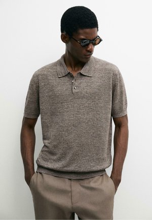 Mężczyzna w brązowym krótkim swetrze polo i beżowych spodniach, z rękami w kieszeniach, patrzący w dół i noszący ciemne okulary przeciwsłoneczne.