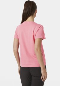 Mujer con cabello largo y oscuro recogido en una coleta, vestida con una camiseta rosa lisa de manga corta y pantalones negros, vista desde atrás.