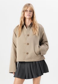 Beige kort jakke med knappelukking, sidelommer og brun krage, parret med et svart plissert skjørt. Glatt tekstur.
