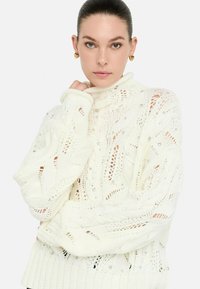 Donna con i capelli raccolti, che indossa orecchini d'oro a forma di cuore e un maglione bianco a trecce decorato con piccole perline.