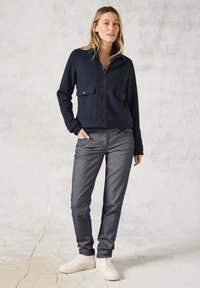 Marine zip-up jack met twee borstzakken, gecombineerd met textuur donkergrijze broek. Model draagt witte sneakers. Achtergrond is neutraal en textuur.