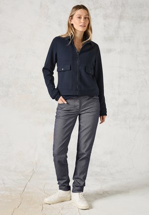 Veste zippée bleu marine avec deux poches poitrine, accompagnée de pantalons texturés gris foncé. Le modèle porte des baskets blanches. L'arrière-plan est neutre et texturé.