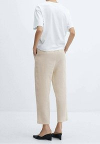 Osoba v béžových zkrácených kalhotách, bílé košili s krátkým rukávem a černých špalíčkových botách, stojící s rukama v kapsách, otočená zády.