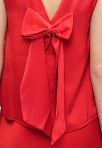 Červený vrchný diel bez rukávov s hlbokým V-výstrihom na chrbte, zdobený veľkou mašľou pri výstrihu a splývavým materiálom, ktorý vytvára jemnú textúru.