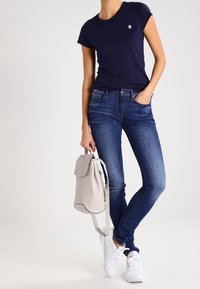 Mörkblå tajt t-shirt, medelblå skinny jeans, vita sneakers och en ljusgrå ryggsäck, stående på en neutral bakgrund.