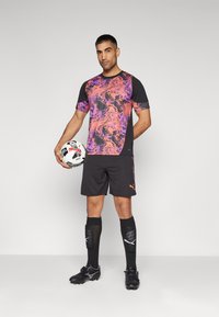 Puma sportfelszerelés, amely egy fekete pólóból áll, élénk lila és narancssárga márványos mintákkal, fekete rövidnadrágból, fekete zoknikból kiemelésekkel és focilabdából.