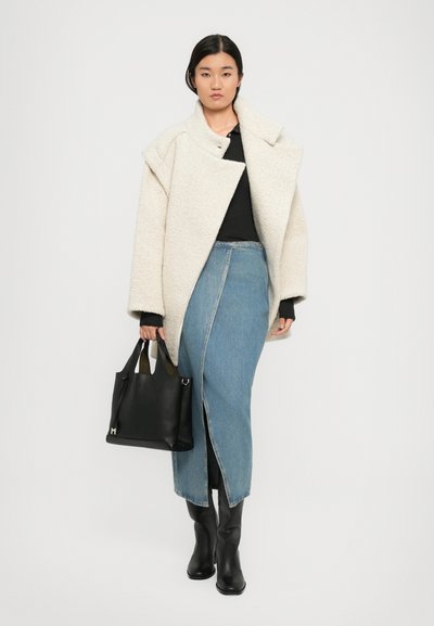 Palton oversized din lână crem, bluză neagră, fustă din denim cu fente adânci, cizme negre din piele până la genunchi și o geantă neagră cu două mânere.