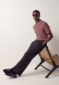 Bordeauxrode geribbelde poloshirt, zwarte broek en glanzende zwarte loafers. Model draagt donkere zonnebril en zit op een gevlochten stoel met een zwart frame.