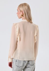 Blusa in chiffon beige con dettagli arricciati sulle spalle, maniche lunghe e polsini con bottoni. Il tessuto ha una leggerezza e una trama fluida.