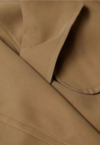 Brauner Stoff mit glatter Textur, der gefaltete Abschnitte und sichtbare Nähdetails aufweist und ein weiches sowie strukturiertes Design zeigt.