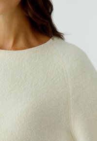 Close-up van een vrouw die een zachte, roomkleurige fuzzy sweater draagt met een ronde halslijn en raglanmouwdetails.