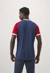 Maglia sportiva con corpo blu navy, maniche raglan rosse con un design testurizzato e rifiniture bianche sul collo e sulle maniche.