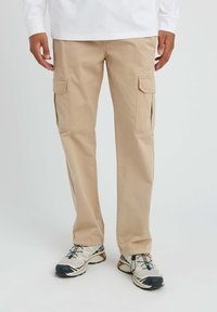 Un pantalon cargo beige avec deux poches latérales, fabriqué en coton. Le design présente des accents cousus et une coupe décontractée, associé à des baskets grises.