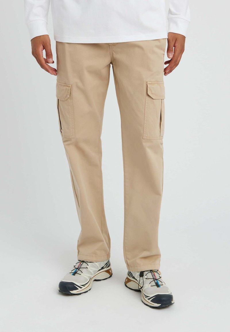 Un pantalon cargo beige avec deux poches latérales, fabriqué en coton. Le design présente des accents cousus et une coupe décontractée, associé à des baskets grises.