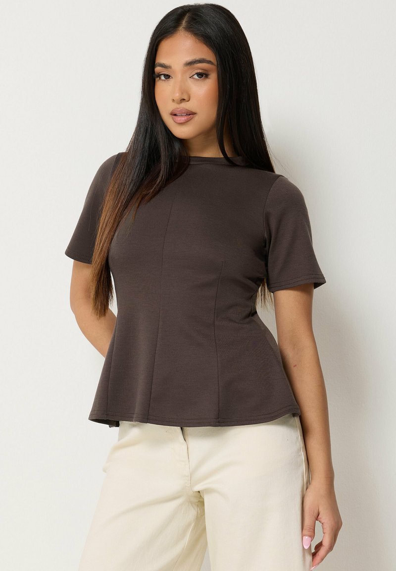 Femme aux longs cheveux foncés portant un haut ajusté à manches courtes marron foncé et un pantalon crème clair sur un fond uni.
