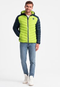 Vaatteita peittävä takki, jossa on kirkkaan lime vihreä etuosa ja tumma laivastonsininen hihnat, sekä vetoketju, huppu ja teksturoitu viisto kuvio.