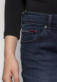 Tmavě modré džíny z denimu mají klasický design s pěti kapsami a viditelným ozdobným prvkem z červeného a bílého prošívání. Detailní záběr na pas.