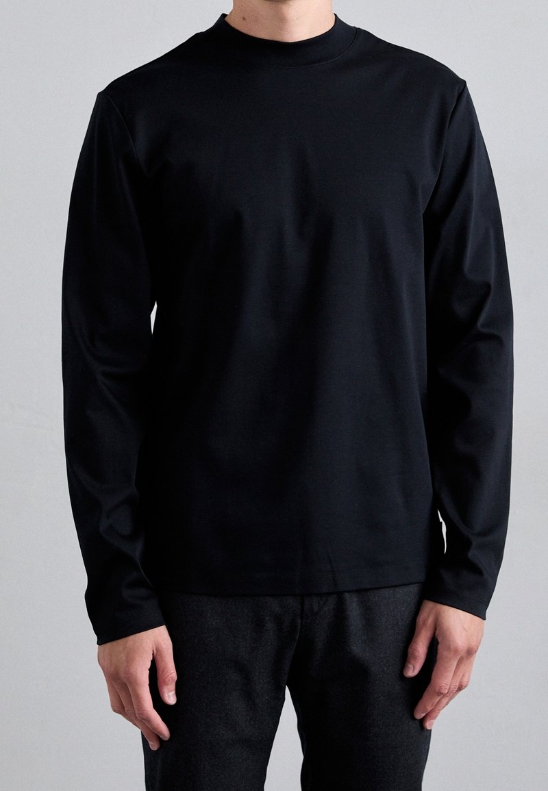 Svart långärmad skjorta i mjukt tyg, med rund halsringning och figursydda ärmar. Kombinerad med mörka texturerade byxor.