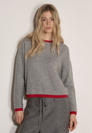 Joven mujer con suéter gris con ribete rojo y pantalones grises a juego con cordón, de pie frente a un fondo claro liso.