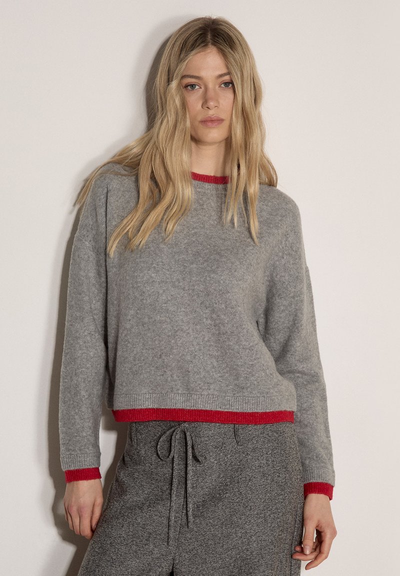 Jeune femme en pull gris avec bordure rouge et pantalon gris à cordon assorti, debout devant un fond clair uni.