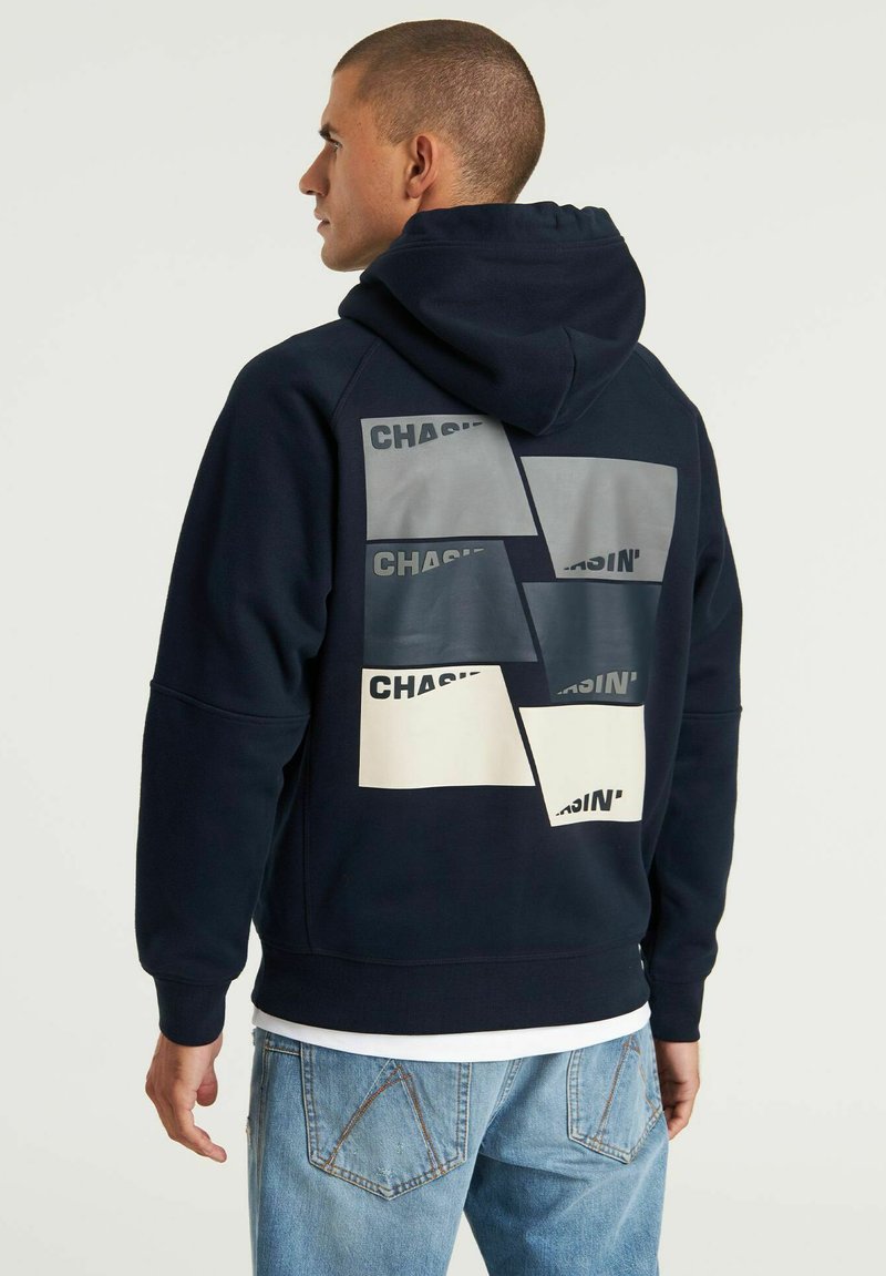 Felpa con cappuccio blu navy con tasche anteriori. Il retro presenta motivi rettangolari sovrapposti in grigio e crema con il testo nero "CHASIN'."