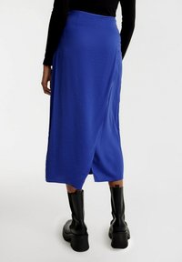 Blauwe midi-rok met een gladde textuur, voorzien van een split aan de achterkant en een aansluitende tailleband. Gecombineerd met zwarte, kniehoge platformlaarzen.