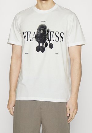 Mann trägt weißes T-Shirt mit schwarzer Pudel-Grafik und den Schriftzügen „FEARLESS“, „HUGO“ und „Since 1993“, kombiniert mit beigen Hosen.
