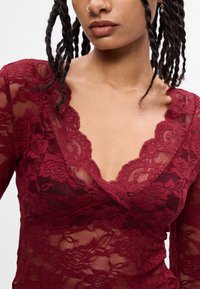 Top de renda burgundy com decote em V, mangas longas e padrões florais. Material transparente com bordas trabalhadas ao longo do decote.