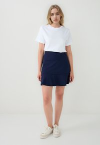 Vrouw die tegen een effen witte achtergrond staat, gekleed in een wit T-shirt, een marineblauwe ruchesrok en witte sneakers.